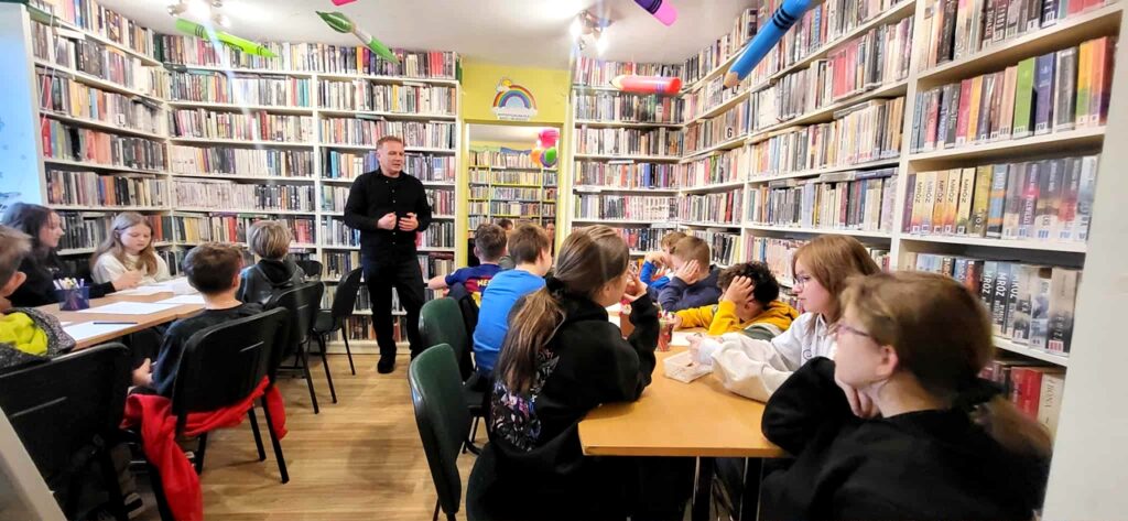 na zdjęciu widać dzieci w sali bibliotecznej siedzące przy stole oraz mówiącego do nich mężczyznę