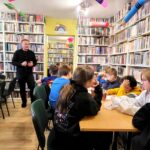 na zdjęciu widać dzieci w sali bibliotecznej siedzące przy stole oraz mówiącego do nich mężczyznę