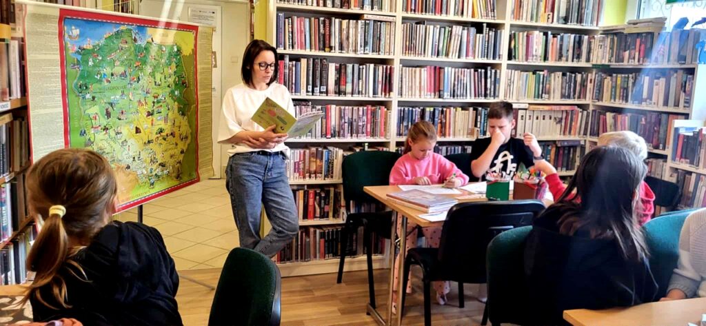 Na zdjęciu widać salę biblioteczną i dzieci siedzące przy stole oraz czytającą im kobietę.