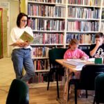 Na zdjęciu widać salę biblioteczną i dzieci siedzące przy stole oraz czytającą im kobietę.