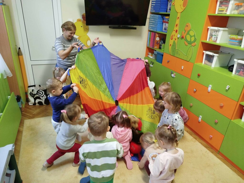 Dzieci wraz z pania bibliotekarką bawia się hustą clanza. Wokół kolorowe mebelki .