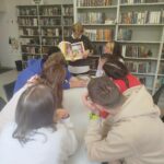 W sali bibliotecznej przy stole siedza kobiety i mężczyźni. W przodzie przed nimi stoi pani bibliotekarka i pokazuje im kolorowa ksiazkę . Za nia w tyle regały z ksiażkami.