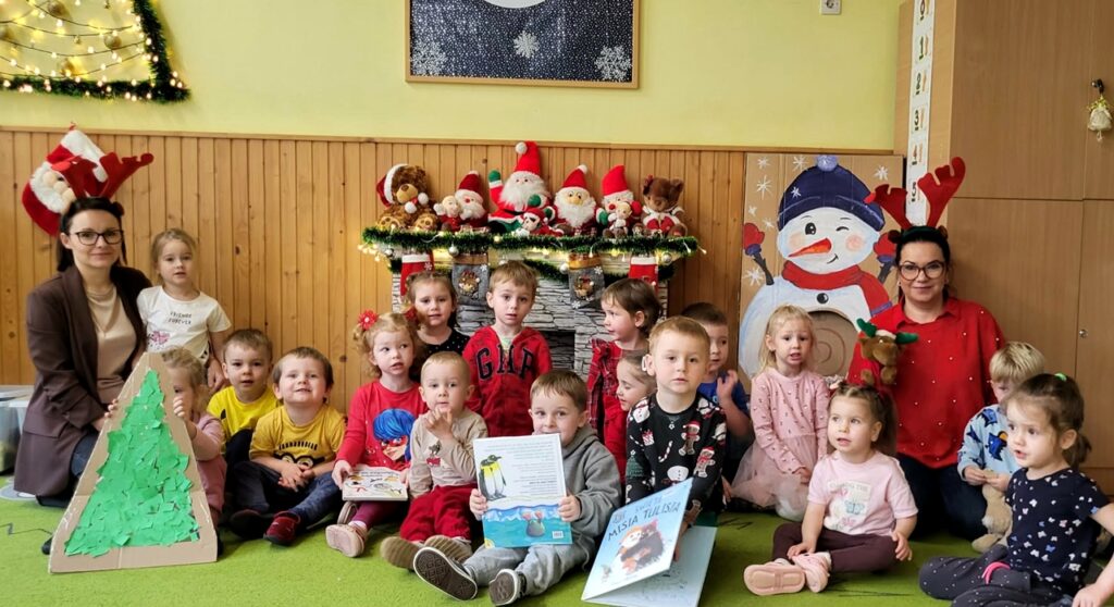 Na tle ściany z Mikołajami siedza przedszkolaki wraz z paniami bibliotekarkami