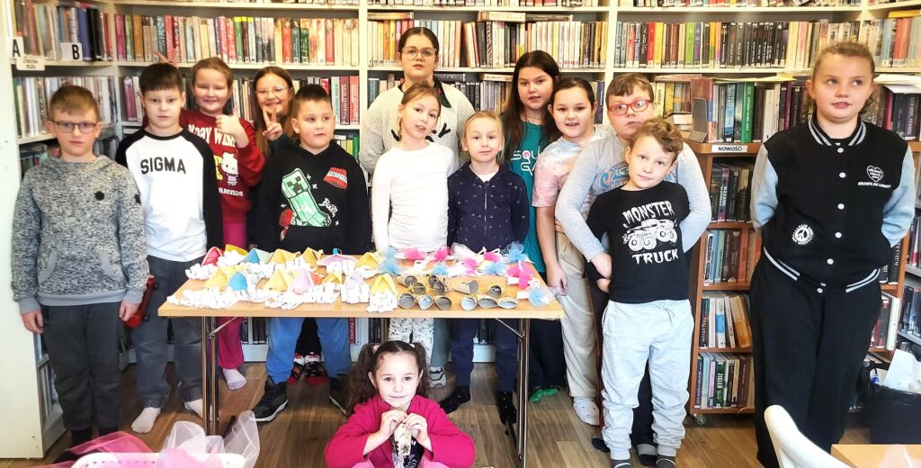 W sali bibliotecznej stoją dzieci za nimi regały z książkami, natomiast przed nimi na stole prace plastyczne wykonane przez ni