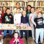 W sali bibliotecznej stoją dzieci za nimi regały z książkami, natomiast przed nimi na stole prace plastyczne wykonane przez ni