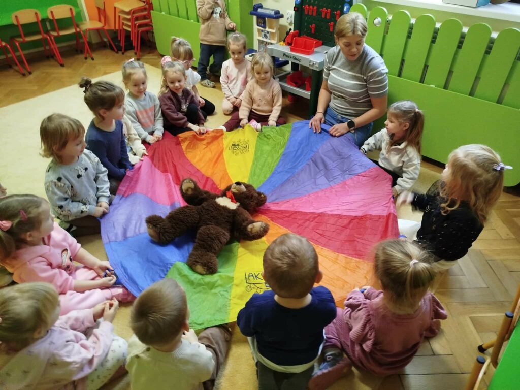 na zdjęciu dzieci siedza w kółeczku na podłodze wraz z pania bibliotekrką. W środku husta clanza a na niej mis pluszowy.