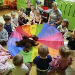 na zdjęciu dzieci siedza w kółeczku na podłodze wraz z pania bibliotekrką. W środku husta clanza a na niej mis pluszowy.