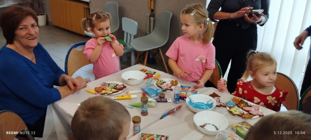 Na zdjęciu dziewczynki siedza wraz ze starsza panią i uczą się dekorować pierniczki .