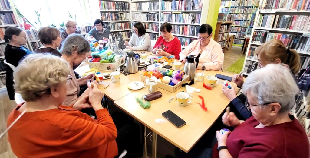 Przy stole w sali bibliotecznej wokół stołu siedza panie seniorki, które uczestniczą w zajęciach plastycznych podczas których wykonuja ozdoby. Wokół regały z książkami .