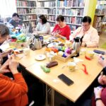 Przy stole w sali bibliotecznej wokół stołu siedza panie seniorki, które uczestniczą w zajęciach plastycznych podczas których wykonuja ozdoby. Wokół regały z książkami .