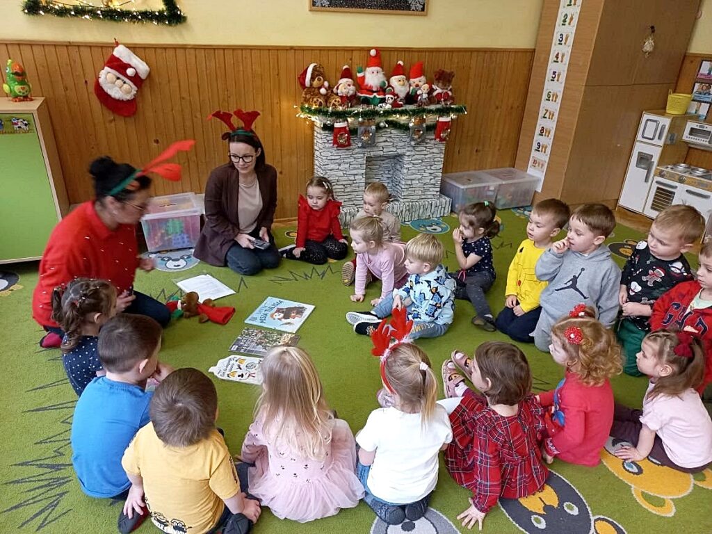 Na sielonym dywanie w sali przrdszkolnej siedza dzieci wraz z paniami bibliotekarkami, które pokazują dzioeciom książeczki kolorowe.