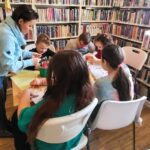 Przy stole w sali bibliotecznej siadzi 5 dzieci w wieku szkolnym przy nich stoi pani bibliotekarka i pokazuje