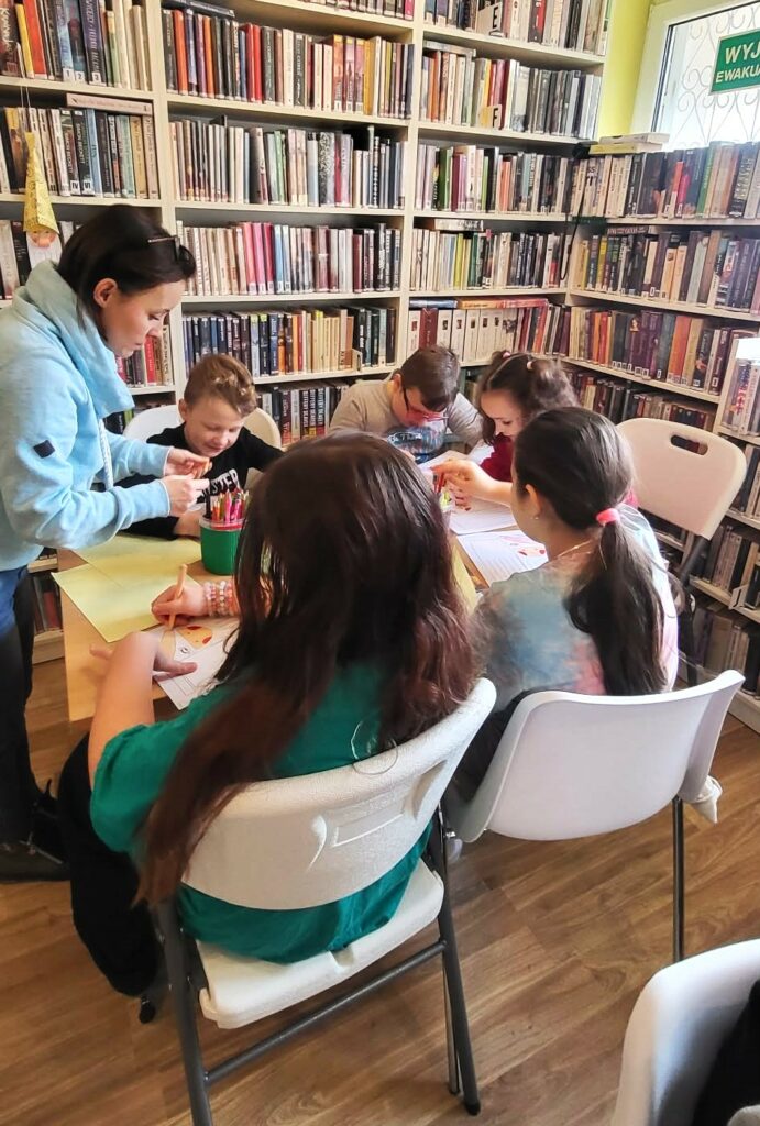 Przy stole w sali bibliotecznej siadzi 5 dzieci w wieku szkolnym przy nich stoi pani bibliotekarka i pokazuje