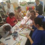 W sali bibliotecznej przy stole siedza kobiety i mężczyźni. Wykonuja kolorowe prace plastyczne. Na stole bardzo dużo materiałów kolorowych ,, kredek, flamastrów.