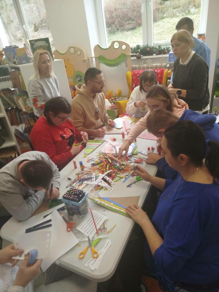 W sali bibliotecznej przy stole siedza kobiety i mężczyźni. Wykonuja kolorowe prace plastyczne. Na stole bardzo dużo materiałów kolorowych ,, kredek, flamastrów.