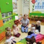 na zdjęciu dzieci siedzą na dywanie w sali przedszkolnej, Wokó nich siedzi pani bibliotekarka, która trzyma w ręku książeczje i pokazuje dzieciom ilustrację.