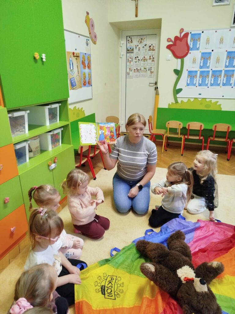 na zdjęciu dzieci siedzą na dywanie w sali przedszkolnej, Wokó nich siedzi pani bibliotekarka, która trzyma w ręku książeczje i pokazuje dzieciom ilustrację.