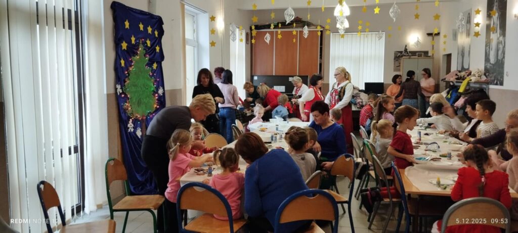 Na zdjęciu dwa długie stoły przy których siedzą dzieci i ozdabiaja pierniczki . Wokół dorosłe panie, które ucza i pomagają dzieciom dekorować.