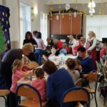 Na zdjęciu dwa długie stoły przy których siedzą dzieci i ozdabiaja pierniczki . Wokół dorosłe panie, które ucza i pomagają dzieciom dekorować.