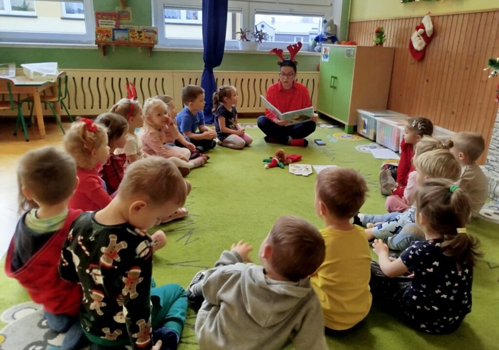 W sali przedszkolnej na zielonym dywanie siedzą przedszkolaki wraz z pania bibliotekarką, która czyta im bajkę.