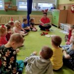 W sali przedszkolnej na zielonym dywanie siedzą przedszkolaki wraz z pania bibliotekarką, która czyta im bajkę.