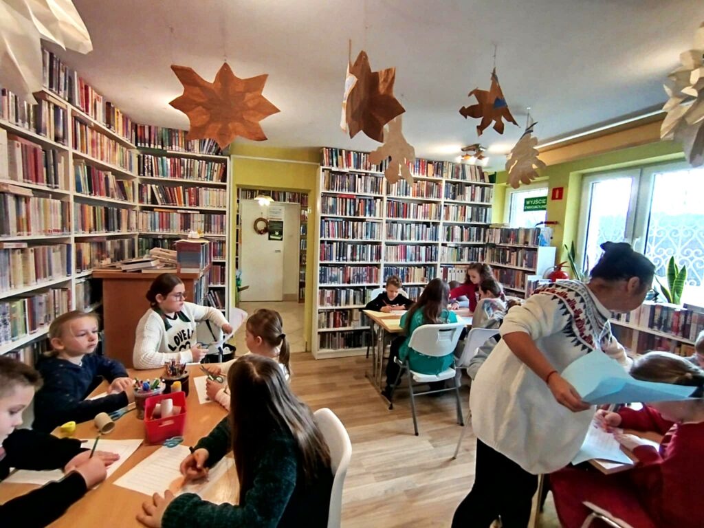 Na zdjęciu sala biblioteczna a w niej dwa stoł przy których siedzi 10 uczestników warsztatów sobotnich. Wykonuja prace plastyczne. na stole kredki