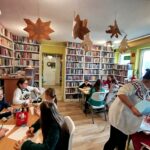 Na zdjęciu sala biblioteczna a w niej dwa stoł przy których siedzi 10 uczestników warsztatów sobotnich. Wykonuja prace plastyczne. na stole kredki
