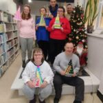 Na zdjęciu w sali bibliotecznej na podwyzszeniu obok choinki stoją trzy kobiety i dwóch mężczyzn. w przodzie siedzi mężczyzna i kobieta.