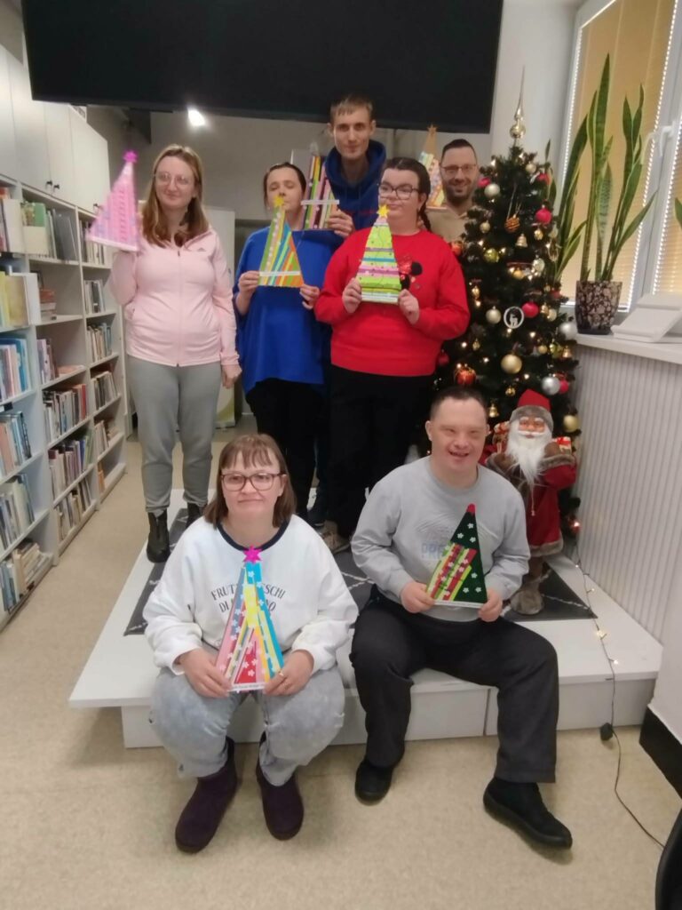 Na zdjęciu w sali bibliotecznej na podwyzszeniu obok choinki stoją trzy kobiety i dwóch mężczyzn. w przodzie siedzi mężczyzna i kobieta.