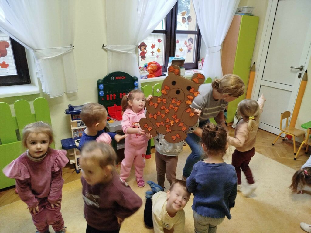 Na zdjęciu grupa dzieci trzyma w tęce kartonowego kolorowego misia. Wokół okna i kolorowe zabawki.