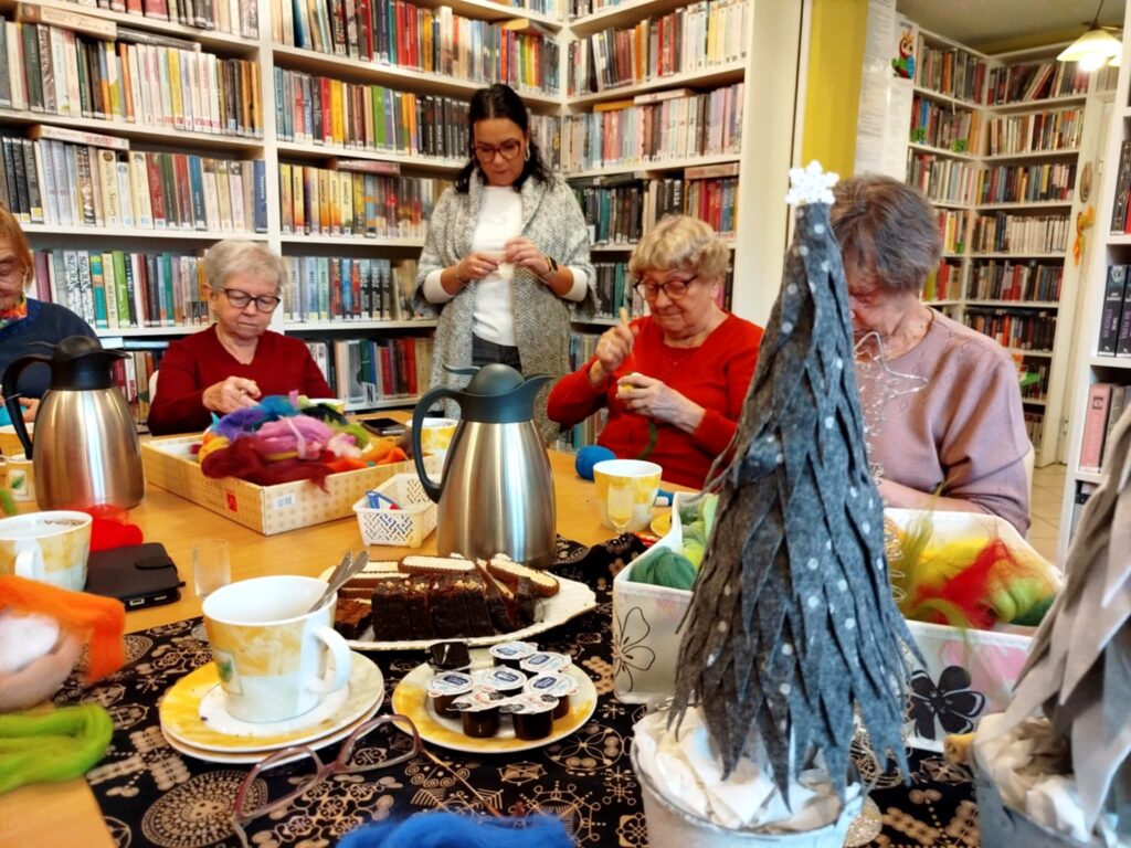 Na zdjęciu w sali bibliotecznej przy stole siedza Panie seniorki i wykonują prace plastyczne . Wokół regały z książkami