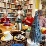 Na zdjęciu w sali bibliotecznej przy stole siedza Panie seniorki i wykonują prace plastyczne . Wokół regały z książkami