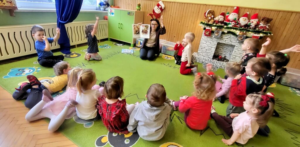 Nazzielonym dywanie siedza przedszkolaki w kółeczku . Siedzi z nimi równiez pani bibliotekarka, która pokazuje kolorowa bajkę. Za nimi na półce siedem maskotek Mikołajów .