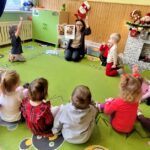 Nazzielonym dywanie siedza przedszkolaki w kółeczku . Siedzi z nimi równiez pani bibliotekarka, która pokazuje kolorowa bajkę. Za nimi na półce siedem maskotek Mikołajów .