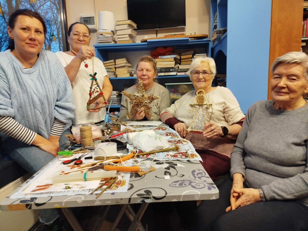 Przy stole siedza trzy kobiety, dwie stoją . W ręku trzymają ozdoby świąteczne .