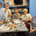 Przy stole siedza trzy kobiety, dwie stoją . W ręku trzymają ozdoby świąteczne .