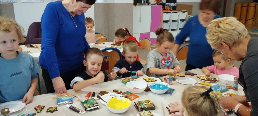 Na zdjęciu dzieci siedza przy stołach i dekorują pierniczki. Przy nich stoja trzy panie seniorki i pomagaja w wykonywaniu prac.
