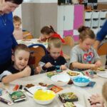 Na zdjęciu dzieci siedza przy stołach i dekorują pierniczki. Przy nich stoja trzy panie seniorki i pomagaja w wykonywaniu prac.