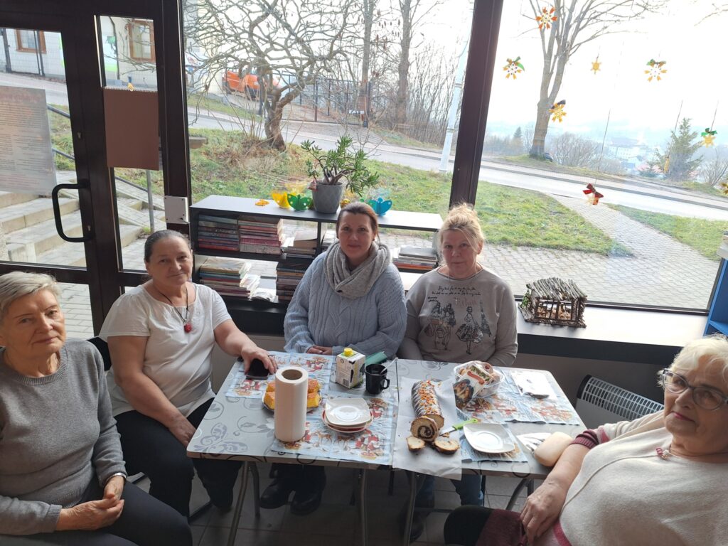 Przy stole siedzi pięc kobiet za nimi w tyle okno za którym widać drogę.