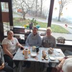 Przy stole siedzi pięc kobiet za nimi w tyle okno za którym widać drogę.