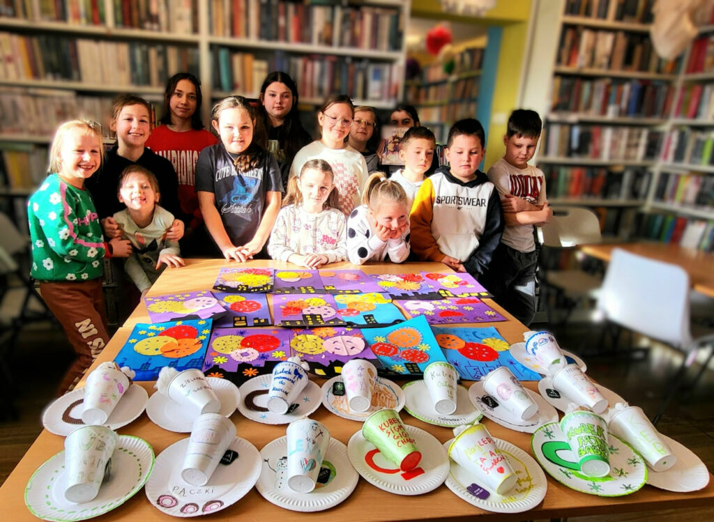 na zdjęciu widać dzieci w sali bibliotecznej stojących za stołem na którym znajdują sie wykonane przez nich prace plastyczne