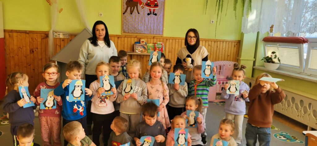 na zdjęciu widać dzieci w sali przedszkolnej trzymające wykonane przez siebie rysunki przedstawiającymi pingwiny
