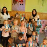 na zdjęciu widać dzieci w sali przedszkolnej trzymające wykonane przez siebie rysunki przedstawiającymi pingwiny