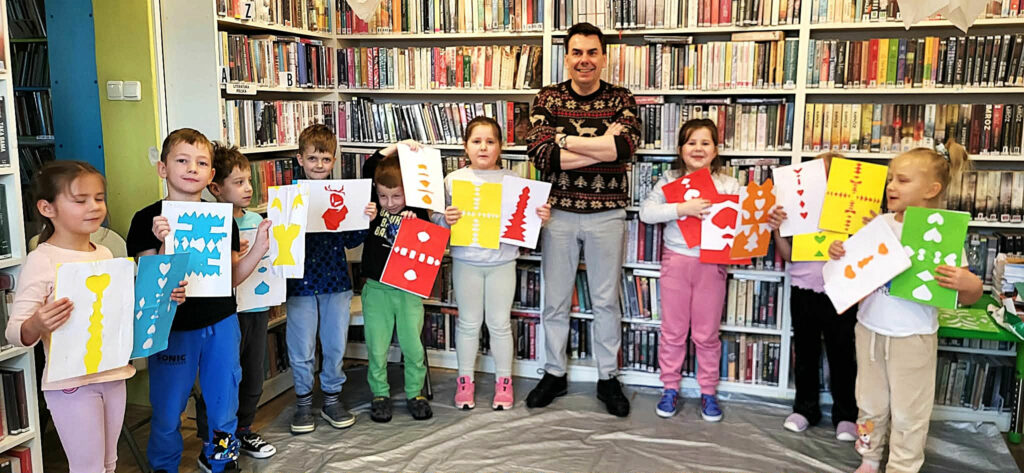 Na zdjęciu dzieci prezentuja prace plastyczne. wśrod nich autor. Za nimi regały z książkami.