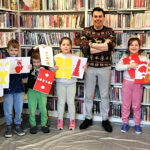 Na zdjęciu dzieci prezentuja prace plastyczne. wśrod nich autor. Za nimi regały z książkami.