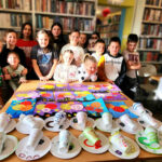 na zdjęciu widać dzieci w sali bibliotecznej stojących za stołem na którym znajdują sie wykonane przez nich prace plastyczne