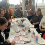 na zdjęciu stół przy którym siedzą kobiety i mężczyźni uczestniczące w warsztatch kreatywno - literackich. Na stole materiały plastyczne potrzebne do warsztatów .