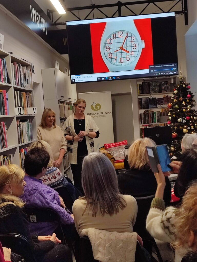 Na zdjęciu wnętrze biblioteki w którym odbywa się spotkanie autorskie z Ewą Chotomską. Na krzesłach tyłem siedza osoby, które przybyły na spotkanie. Z przodu widać kobietę , która przemawia.
