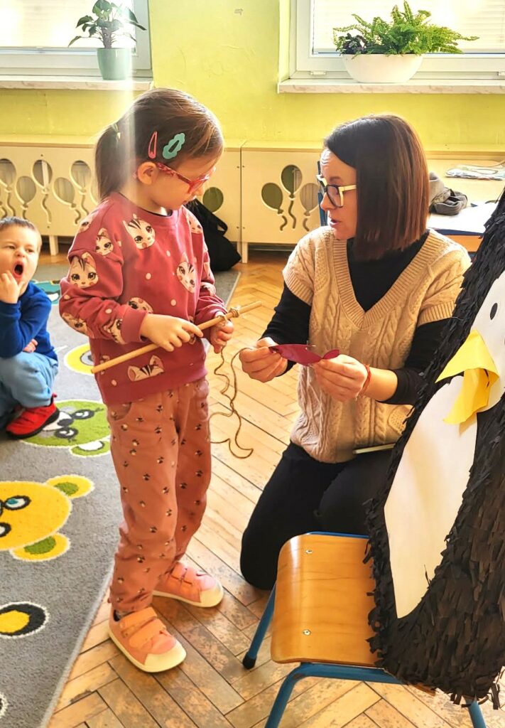 Na zdjęciu pani bibliotekarka kleczy przy krześle przed dziewczynką w wieku przedszkolnym. Pani bibliotekarka tłumaczy ciś dziewczynce.