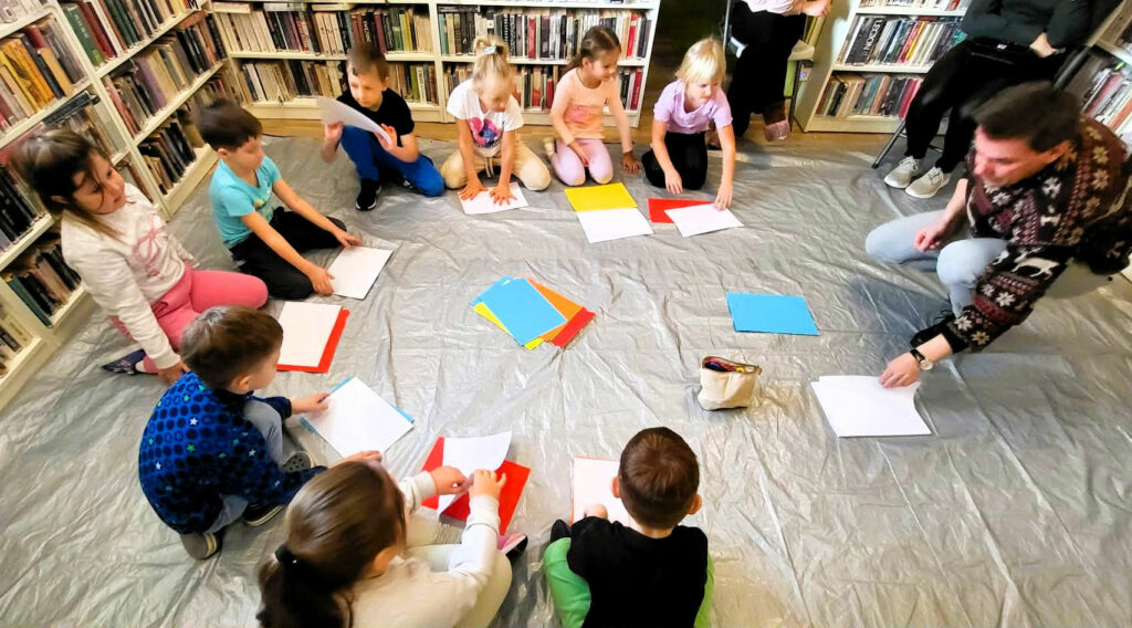 Na zdjęciu dzieci siedzące w kółku na ziemi przed nimi prowadzący . Na ziemi przed kazdym dzieckiem kolorowe kartki A4. Za nimi regały a na nich ksiazki.